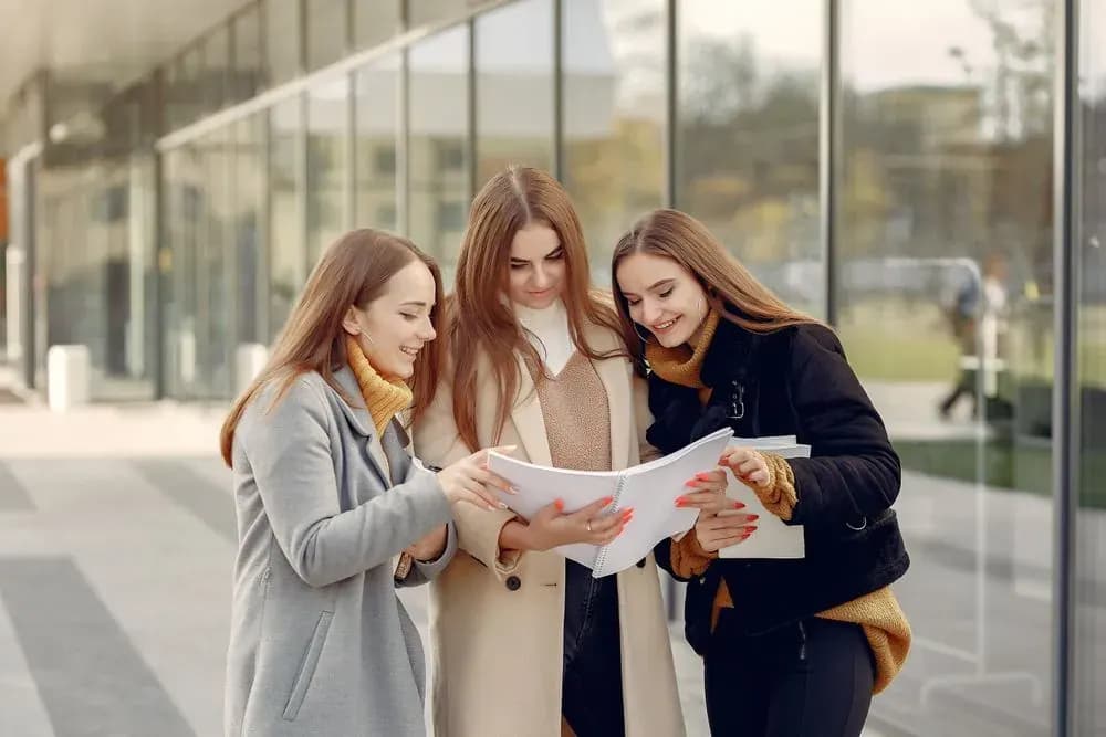 Three students reading a university brochure outside a modern campus building